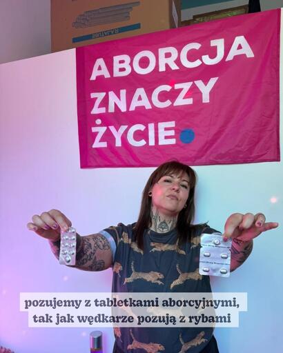 Kobieta pozuje z tabletkami aborcyjnymi na tle różowej flagi z napisem „ABORCJA ZNACZY ŻYCIE”. 

podpis: 
„Pozujemy z tabletkami poronnymi, tak jak rybacy pozują z rybami”.